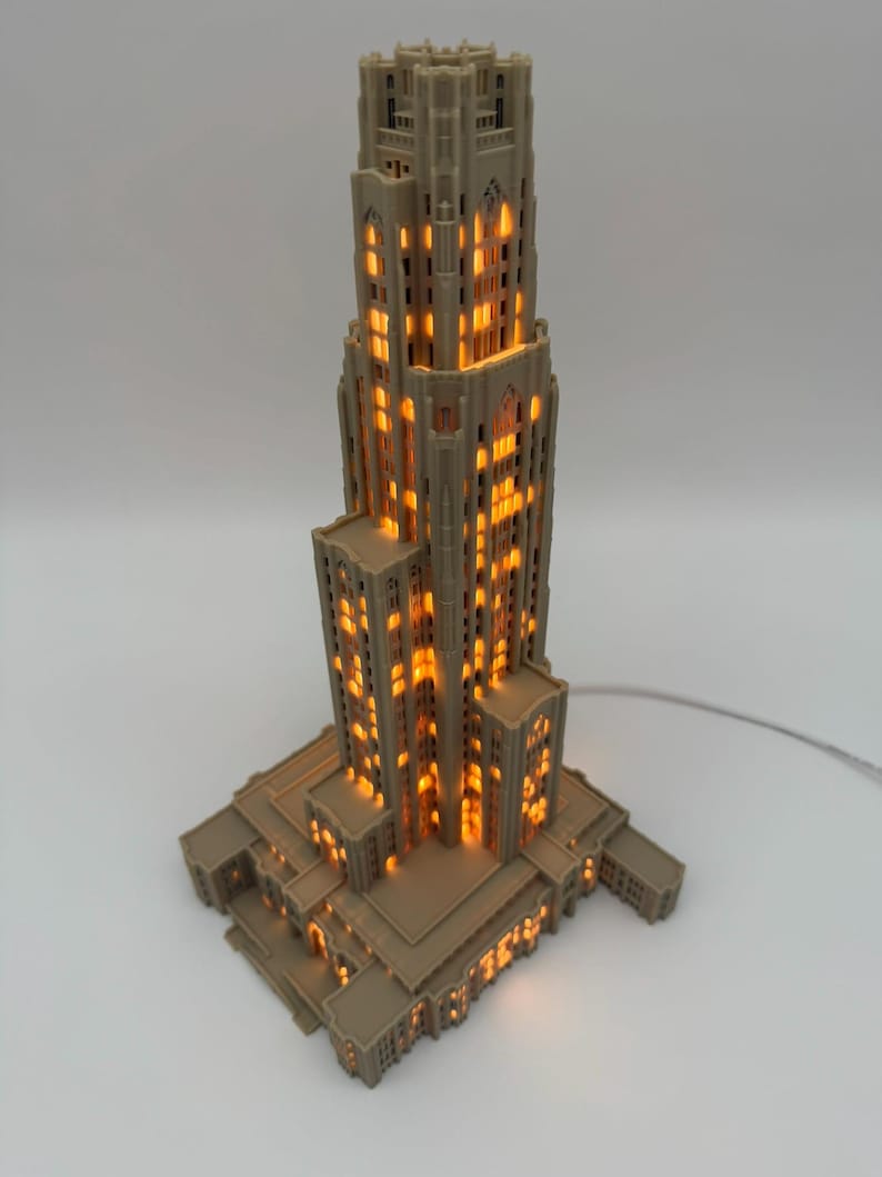 May include: A beige, 3D-printed architectural model of a tall skyscraper with a stepped design. The structure features numerous rectangular windows that emit a warm, glowing orange light from within, creating a realistic illuminated effect.