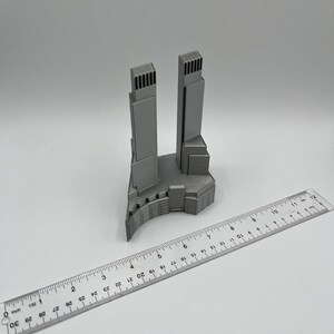 Op de afbeelding: Een 3D-geprint model van een paar wolkenkrabbers met een gebogen basis. Het model is gemaakt van grijs plastic en is ongeveer 10 cm hoog.