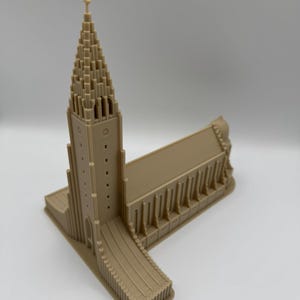 May include: A beige architectural model of a church with a tall, pointed steeple and a rectangular base. The model has detailed features, including windows and a cross atop the steeple. The overall design is symmetrical and detailed.