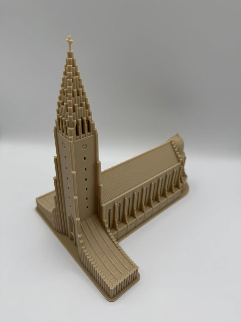 May include: A beige architectural model of a church with a tall, pointed steeple and a rectangular base. The model has detailed features, including windows and a cross atop the steeple. The overall design is symmetrical and detailed.