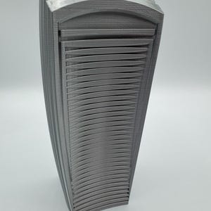 May include: A gray, 3D-printed decorative object with a curved, layered design. The object has a series of horizontal, curved lines that create a textured surface. The overall shape is rectangular with rounded edges, and it stands upright on a white surface.