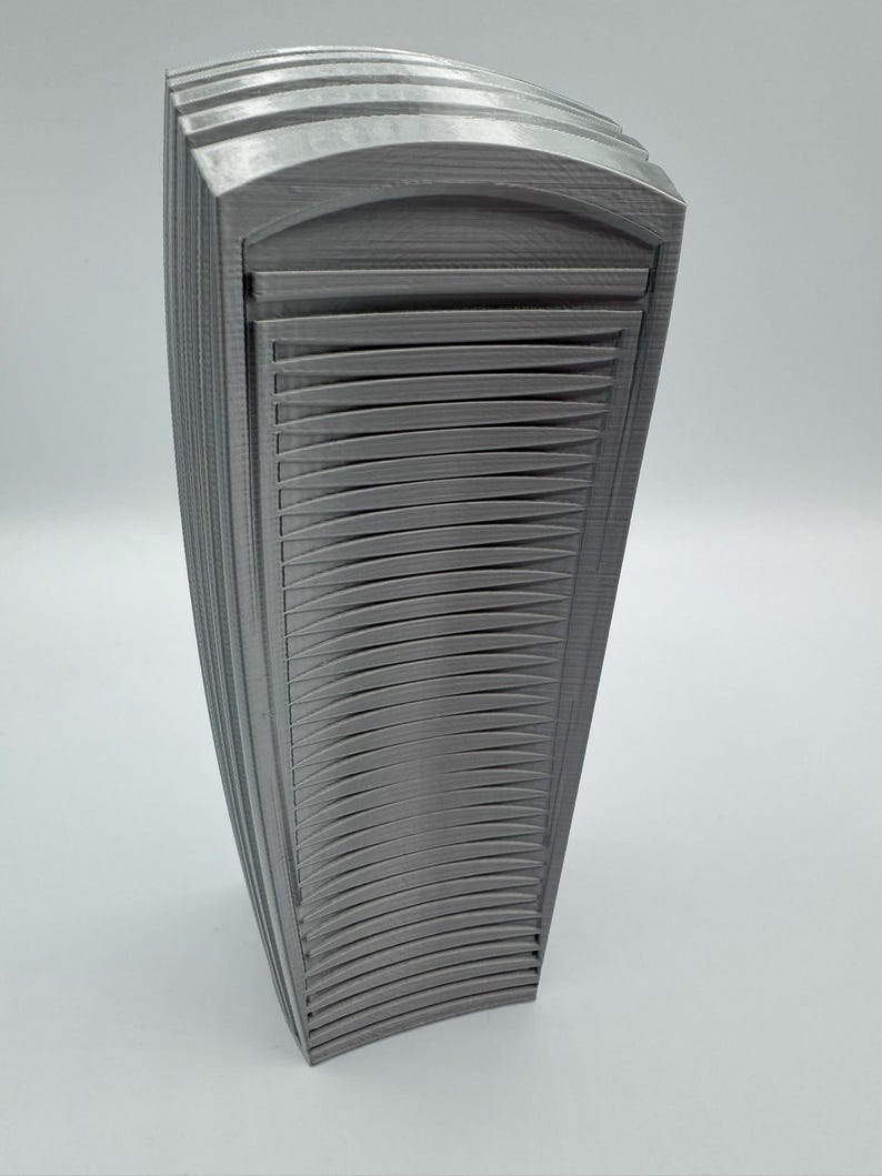May include: A gray, 3D-printed decorative object with a curved, layered design. The object has a series of horizontal, curved lines that create a textured surface. The overall shape is rectangular with rounded edges, and it stands upright on a white surface.