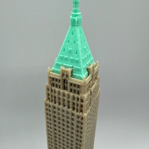 May include: A 3D printed model of a tall building with a green roof and a pointed spire. The building is made of a light brown plastic and has many windows.