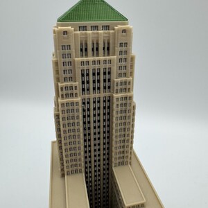 May include: A detailed model of a skyscraper with a beige facade, a green roof, and a small statue at the top. The building has many windows and a tall, slender design, showcasing architectural detail.