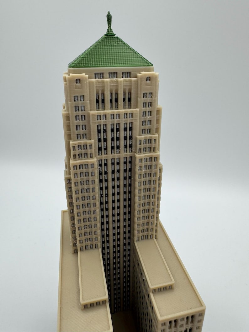 May include: A detailed model of a skyscraper with a beige facade, a green roof, and a small statue at the top. The building has many windows and a tall, slender design, showcasing architectural detail.