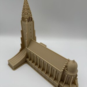 May include: A tan-colored miniature model of a church with a tall, pointed steeple and a cross at the top. The building has multiple arched windows and a domed structure at one end. The model is made of a solid material.