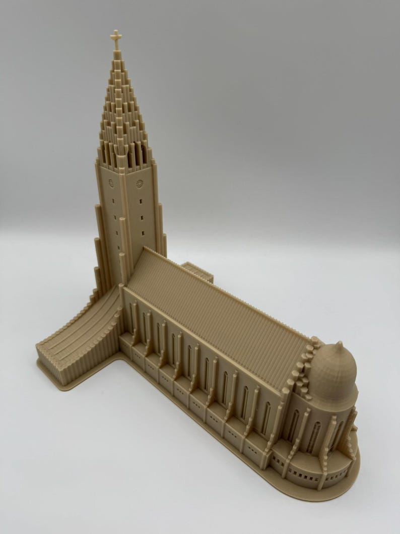 May include: A tan-colored miniature model of a church with a tall, pointed steeple and a cross at the top. The building has multiple arched windows and a domed structure at one end. The model is made of a solid material.