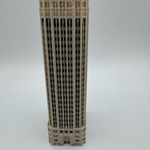 May include: A beige 3D printed architectural model of a skyscraper featuring a rectangular structure with vertical columns, recessed windows, and intricate detailing at the top section. The model displays a uniform light tan color throughout.