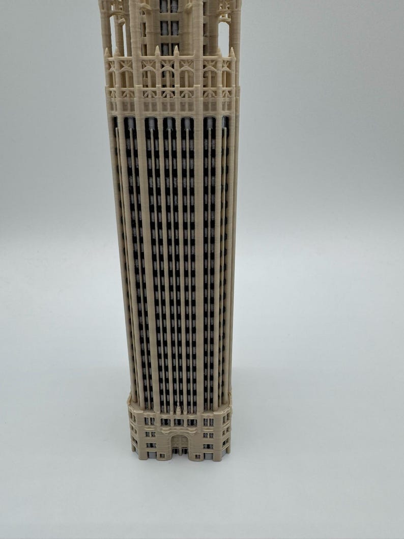 May include: A beige 3D printed architectural model of a skyscraper featuring a rectangular structure with vertical columns, recessed windows, and intricate detailing at the top section. The model displays a uniform light tan color throughout.