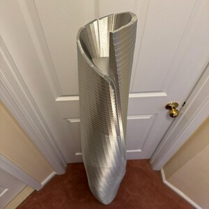 May include: A tall, silver, decorative vase with a unique, twisted design. The vase is made of metal and has a smooth, shiny finish.