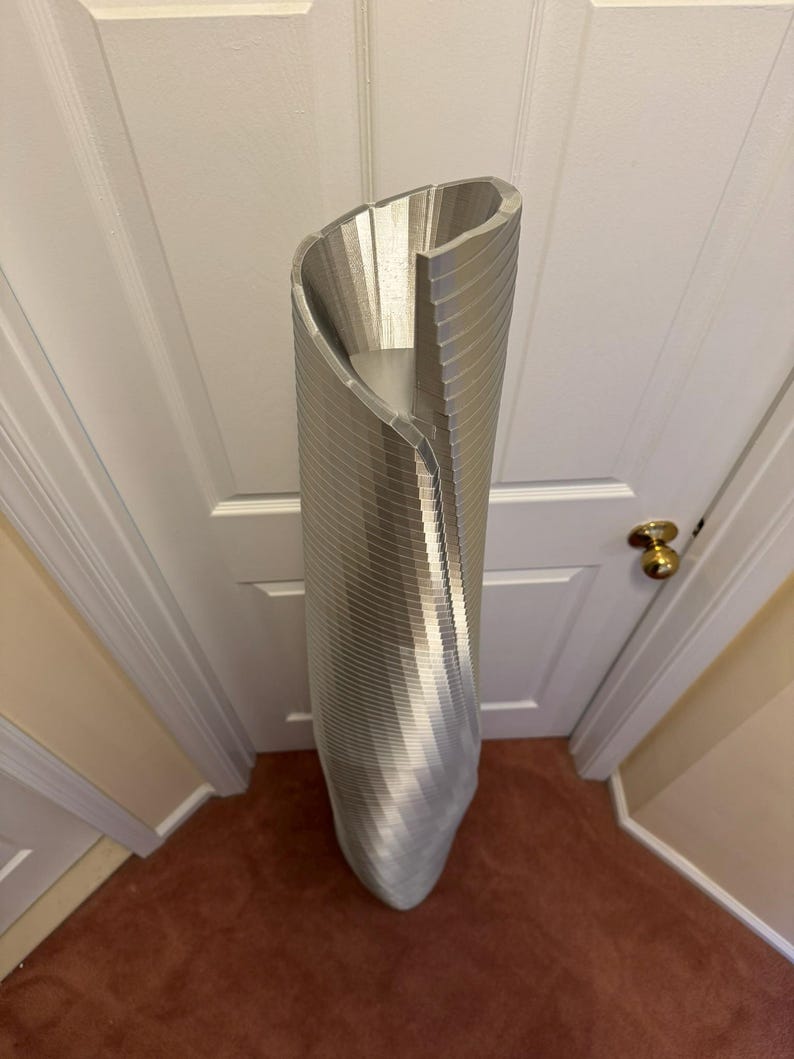 May include: A tall, silver, decorative vase with a unique, twisted design. The vase is made of metal and has a smooth, shiny finish.