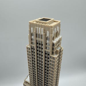 May include: A beige miniature skyscraper model with a detailed facade, featuring numerous windows and architectural elements. The building has a stepped design and a flat roof with a dark square feature. The model is set against a plain background.