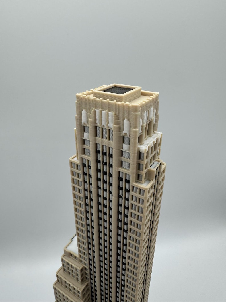 May include: A beige miniature skyscraper model with a detailed facade, featuring numerous windows and architectural elements. The building has a stepped design and a flat roof with a dark square feature. The model is set against a plain background.
