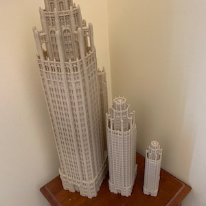May include: Three white 3D printed models of skyscrapers, the largest is about 25 cm tall, the middle is about 15 cm tall, and the smallest is about 10 cm tall. The models are on a wooden table with a ruler.