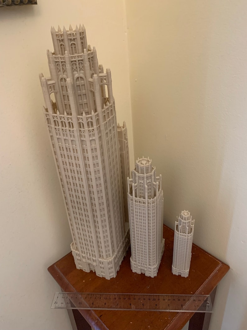 May include: Three white 3D printed models of skyscrapers, the largest is about 25 cm tall, the middle is about 15 cm tall, and the smallest is about 10 cm tall. The models are on a wooden table with a ruler.