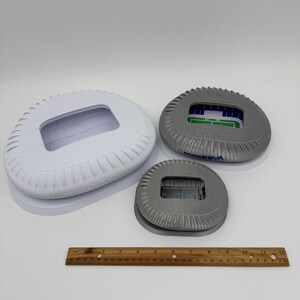 May include: Three stadium models in varying sizes. The largest is white, the medium is gray with green and blue accents, and the smallest is gray. A wooden ruler is placed below the models.