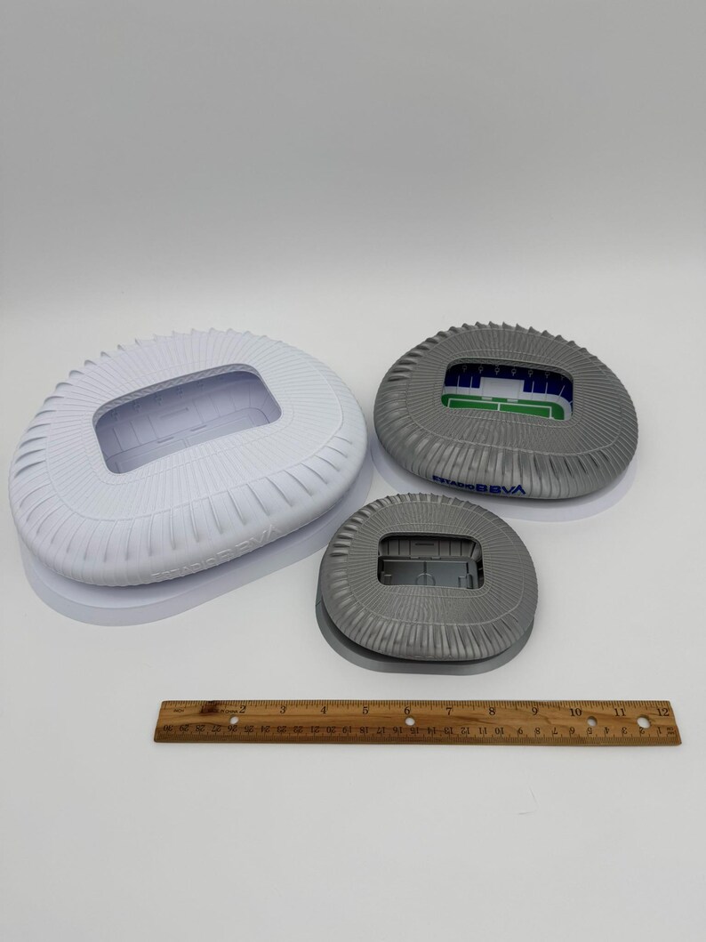 May include: Three stadium models in varying sizes. The largest is white, the medium is gray with green and blue accents, and the smallest is gray. A wooden ruler is placed below the models.