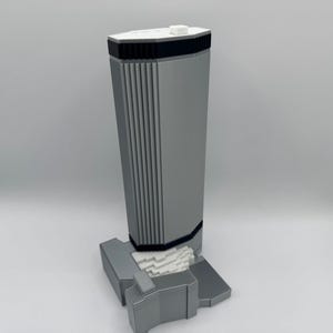 May include: A grey architectural model of a skyscraper. The building has a rectangular shape with vertical lines on the front. The base is a lighter grey with a white, textured section. The top has a black border and a small white square.