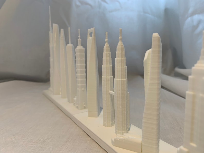 May include: A collection of white 3D-printed miniature skyscraper models, including the Taipei 101 and the Petronas Towers. The buildings are arranged in a row on a white base, showcasing architectural details.