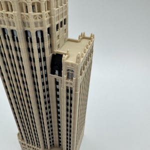 May include: A 3D printed architectural model of a skyscraper featuring a light beige exterior with vertical recessed sections. The structure displays intricate facade details, including multiple window rows, decorative crenellations, and black accents.