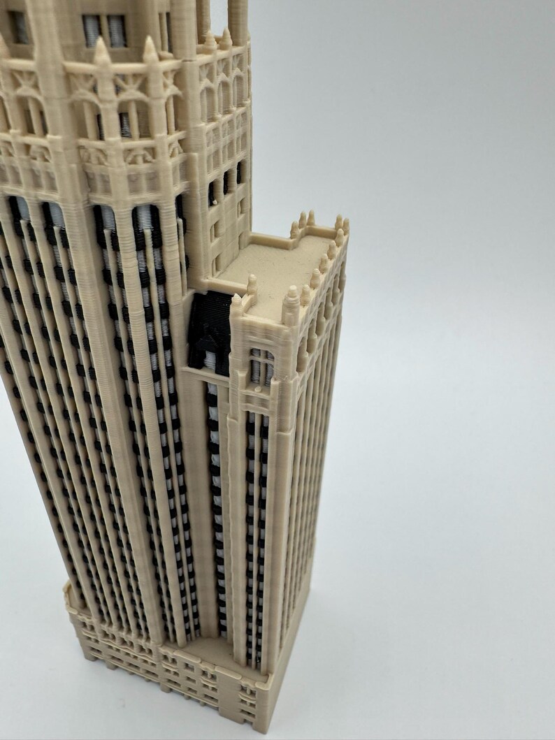 May include: A 3D printed architectural model of a skyscraper featuring a light beige exterior with vertical recessed sections. The structure displays intricate facade details, including multiple window rows, decorative crenellations, and black accents.