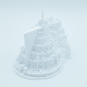 May include: A white 3D printed model of a fantasy city with a tall tower and surrounding buildings. The city is built on a rocky base.