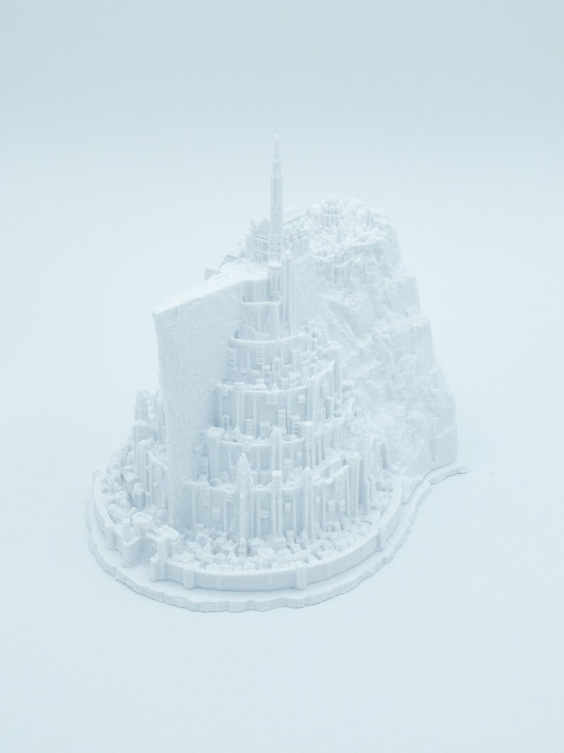 May include: A white 3D printed model of a fantasy city with a tall tower and surrounding buildings. The city is built on a rocky base.