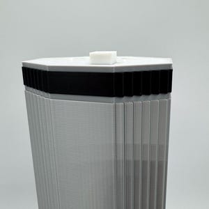 May include: A tall, gray, geometric structure with a hexagonal top. The top is white with a small, white, rectangular block on top. A black band encircles the structure below the top. The sides are vertically ribbed.