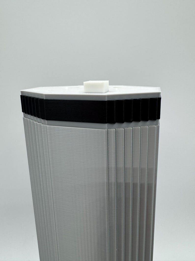 May include: A tall, gray, geometric structure with a hexagonal top. The top is white with a small, white, rectangular block on top. A black band encircles the structure below the top. The sides are vertically ribbed.