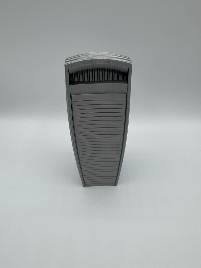 May include: A gray, architectural-style object with a rectangular shape and a slatted design on the front. The top features a series of vertical lines, and the overall appearance is modern and minimalist.
