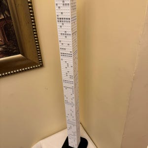 May include: A tall, white model of a skyscraper with a black base. The building has many small, square windows and is set against a beige wall. The model is a detailed representation of a modern high-rise.