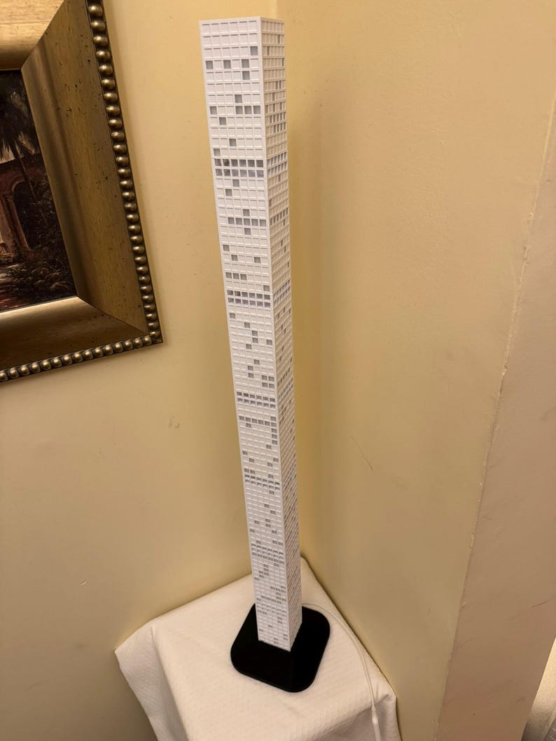 May include: A tall, white model of a skyscraper with a black base. The building has many small, square windows and is set against a beige wall. The model is a detailed representation of a modern high-rise.