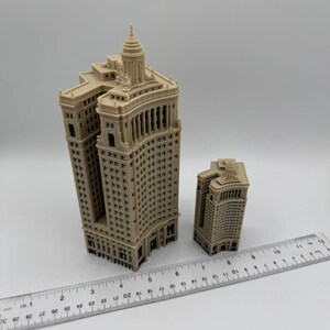 Op de afbeelding: Twee miniatuurmodellen van gebouwen, een groter dan de andere, zijn gemaakt van beige plastic en hebben een gedetailleerd ontwerp. De gebouwen staan op een wit oppervlak met een liniaal ernaast.