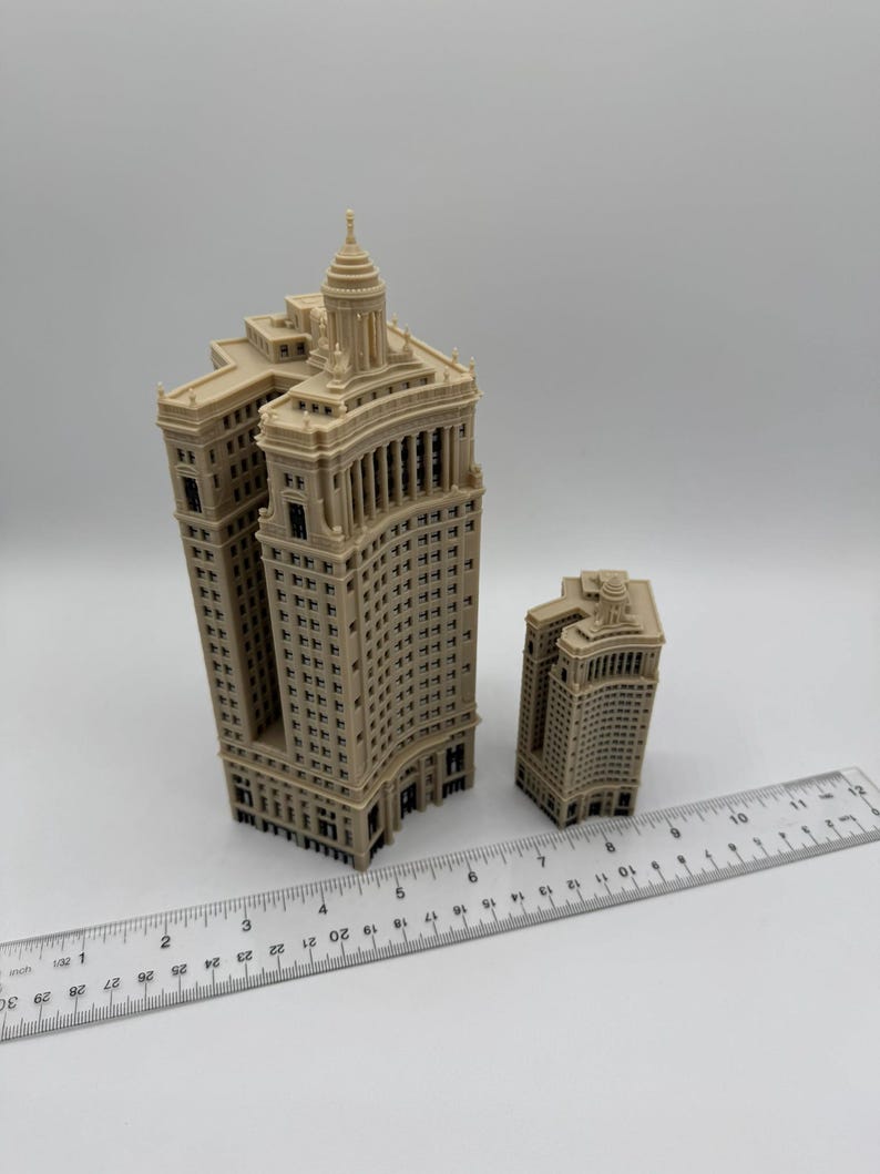 Op de afbeelding: Twee miniatuurmodellen van gebouwen, een groter dan de andere, zijn gemaakt van beige plastic en hebben een gedetailleerd ontwerp. De gebouwen staan op een wit oppervlak met een liniaal ernaast.