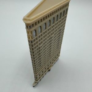 May include: A beige 3D printed model of the Flatiron Building in New York City. The model is detailed with windows and a triangular roof.
