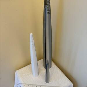 May include: Three silver-colored metal sculptures of different sizes. The sculptures are shaped like tall, slender towers with intricate designs. The sculptures are arranged on a white tablecloth with a ruler for scale.