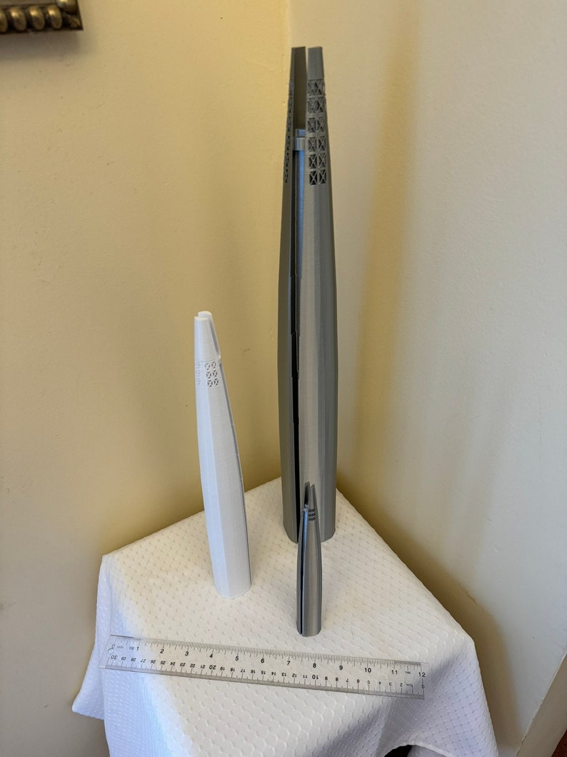May include: Three silver-colored metal sculptures of different sizes. The sculptures are shaped like tall, slender towers with intricate designs. The sculptures are arranged on a white tablecloth with a ruler for scale.