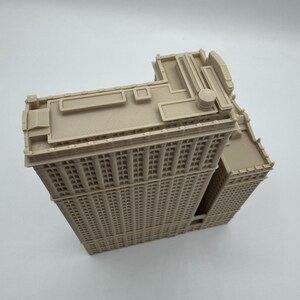 May include: A beige, 3D-printed model of a tall building with many windows. The building has a flatiron shape, with a triangular footprint. The model has detailed architectural features, including a detailed roof and window design. The model is made of a light beige plastic.
