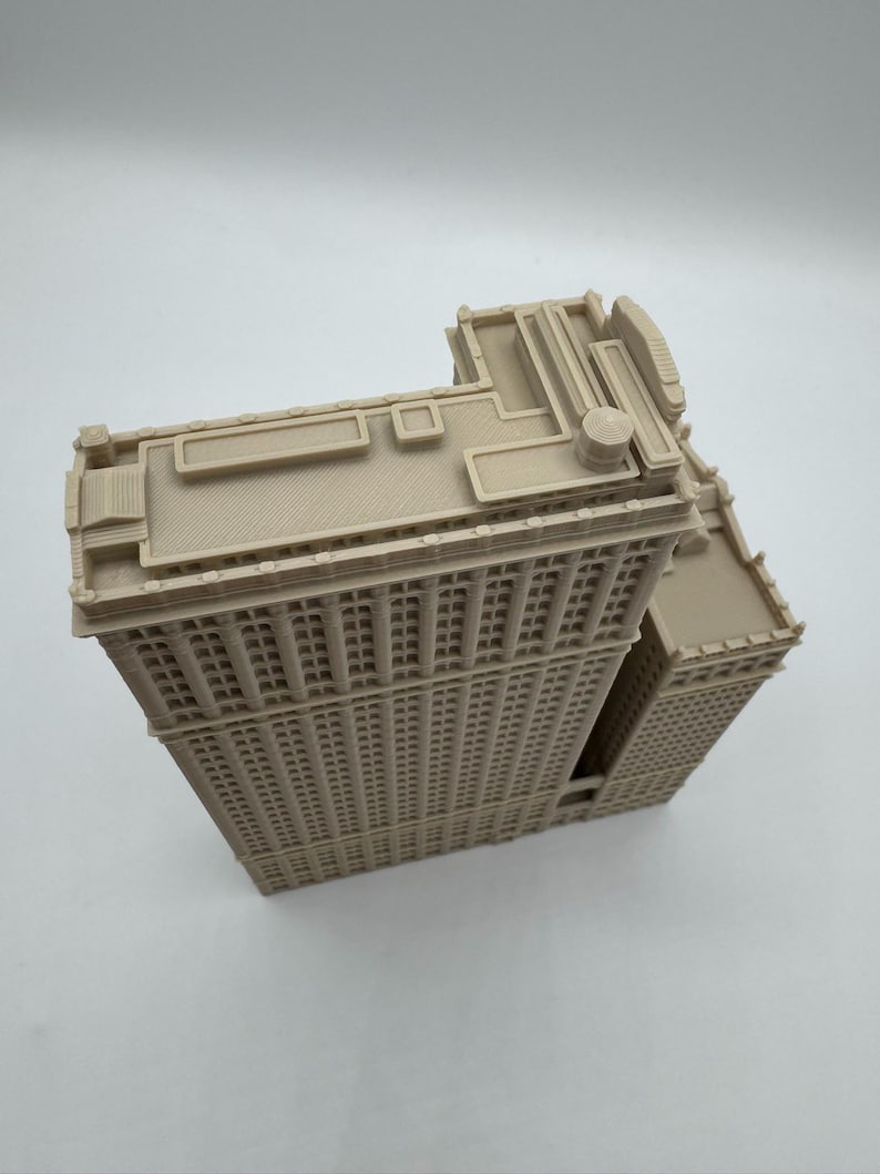 May include: A beige, 3D-printed model of a tall building with many windows. The building has a flatiron shape, with a triangular footprint. The model has detailed architectural features, including a detailed roof and window design. The model is made of a light beige plastic.