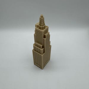 May include: A tan-colored miniature skyscraper model with a detailed, textured facade. The building has a tiered design, with a central tower and multiple levels. The model is standing on a white surface.