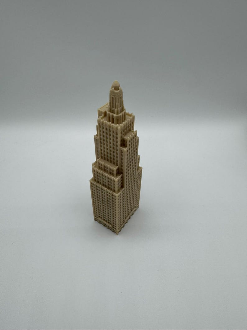 May include: A tan-colored miniature skyscraper model with a detailed, textured facade. The building has a tiered design, with a central tower and multiple levels. The model is standing on a white surface.