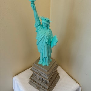 May include: A teal blue 3D printed statue of the Statue of Liberty with a torch raised high. The statue is standing on a brown base with a layered design.