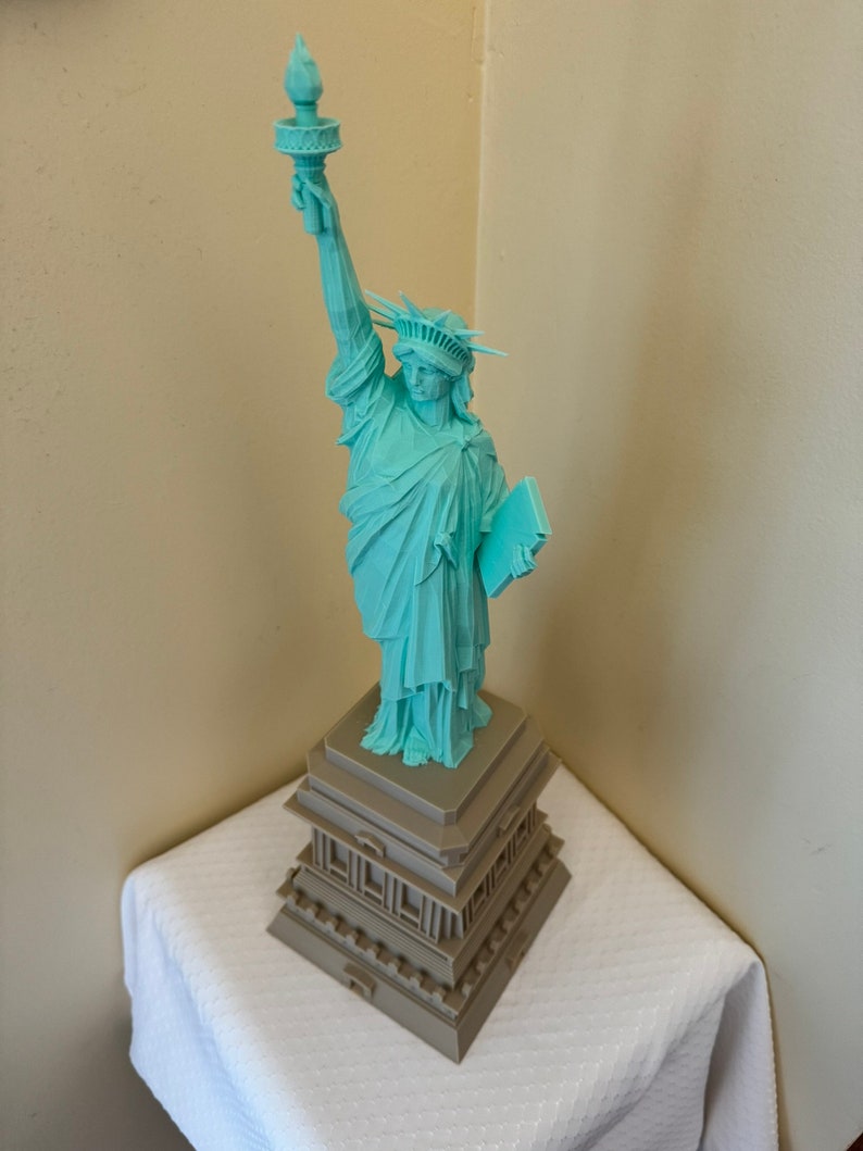 May include: A teal blue 3D printed statue of the Statue of Liberty with a torch raised high. The statue is standing on a brown base with a layered design.