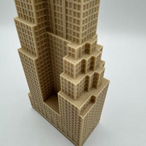 May include: A tan-colored miniature of a skyscraper, featuring a detailed facade with numerous windows. The building has a stepped design, with multiple tiers and a rectangular base. The model is likely a decorative item or architectural replica.