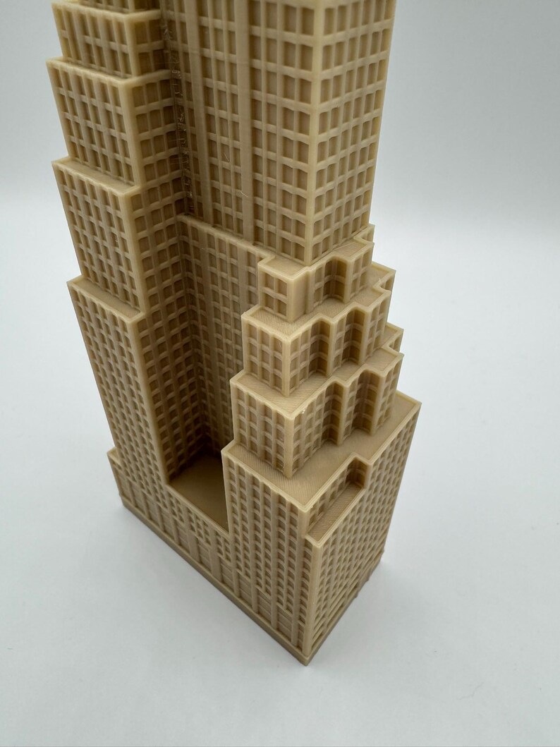 May include: A tan-colored miniature of a skyscraper, featuring a detailed facade with numerous windows. The building has a stepped design, with multiple tiers and a rectangular base. The model is likely a decorative item or architectural replica.