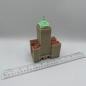 May include: A miniature model of a tall building with a beige facade, red roof accents, and a green-topped spire. A clear ruler with inch markings is placed next to the building, showing its size.