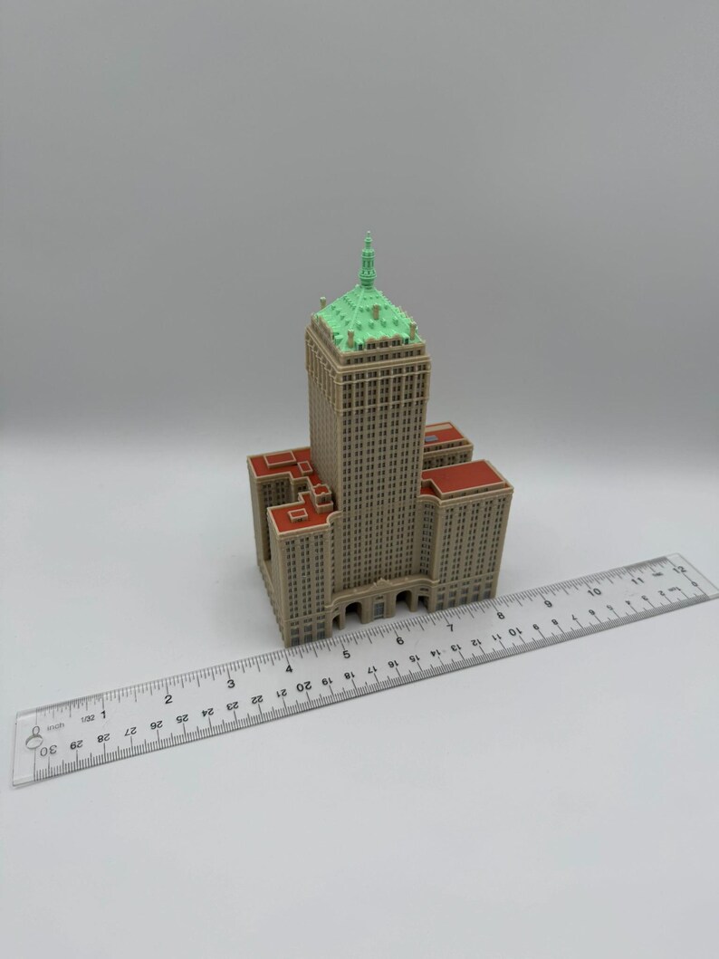 May include: A miniature model of a tall building with a beige facade, red roof accents, and a green-topped spire. A clear ruler with inch markings is placed next to the building, showing its size.
