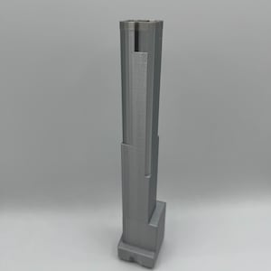 Peut inclure: Un objet gris imprimé en 3D, haut et vertical, au design géométrique étagé. La structure présente une base rectangulaire qui se transforme en une tour plus étroite à plusieurs niveaux, avec des lignes de couche horizontales visibles.