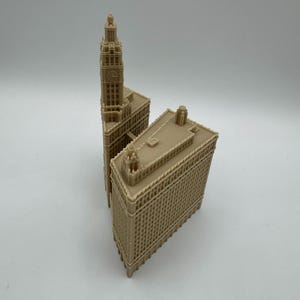 May include: Two beige miniature architectural models of buildings. One model features a tall clock tower, while the other is a rectangular building. Both models have detailed facades and are set against a white background.
