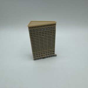 May include: A miniature model of a tall, beige building with many windows. The building has a triangular roof and a detailed facade.
